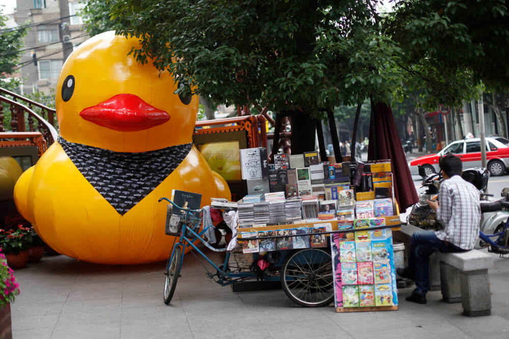 A scaled replica of the "Rubber Duck" by Dutch conceptual artist Florentijn Hofman is seen along a street next to a vendor waiting for customers in Shanghai